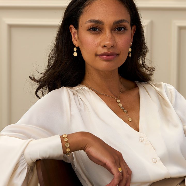 Schmuck für Damen Frau mit braunem Haar, einem weißen Blusenshirt und goldener Schmuck sitzt entspannt.